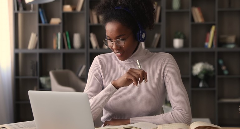 Black woman with glasses, headphones and holding pen, studying in front of laptop and open books